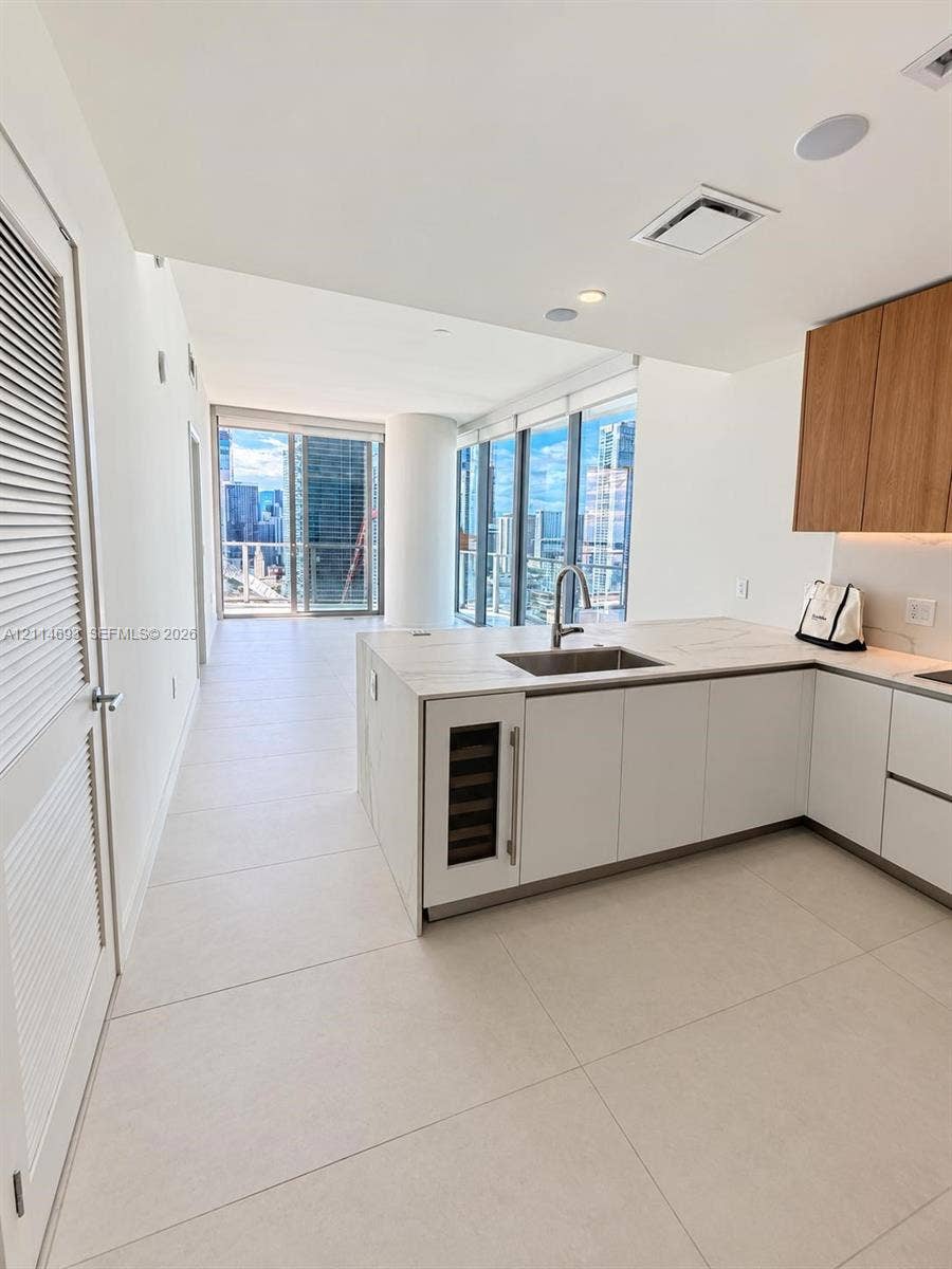 Kitchen featuring large-format tile flooring, a stone-finish island with an integrated wine refrigerator, custom wood-tone upper cabinetry, and floor-to-ceiling glass windows