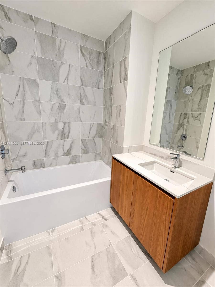 Bathroom featuring a wood-finish vanity with a white countertop and integrated sink, a large wall-mounted mirror, a tub-and-shower combination, and full-height stone-look tile surrounds