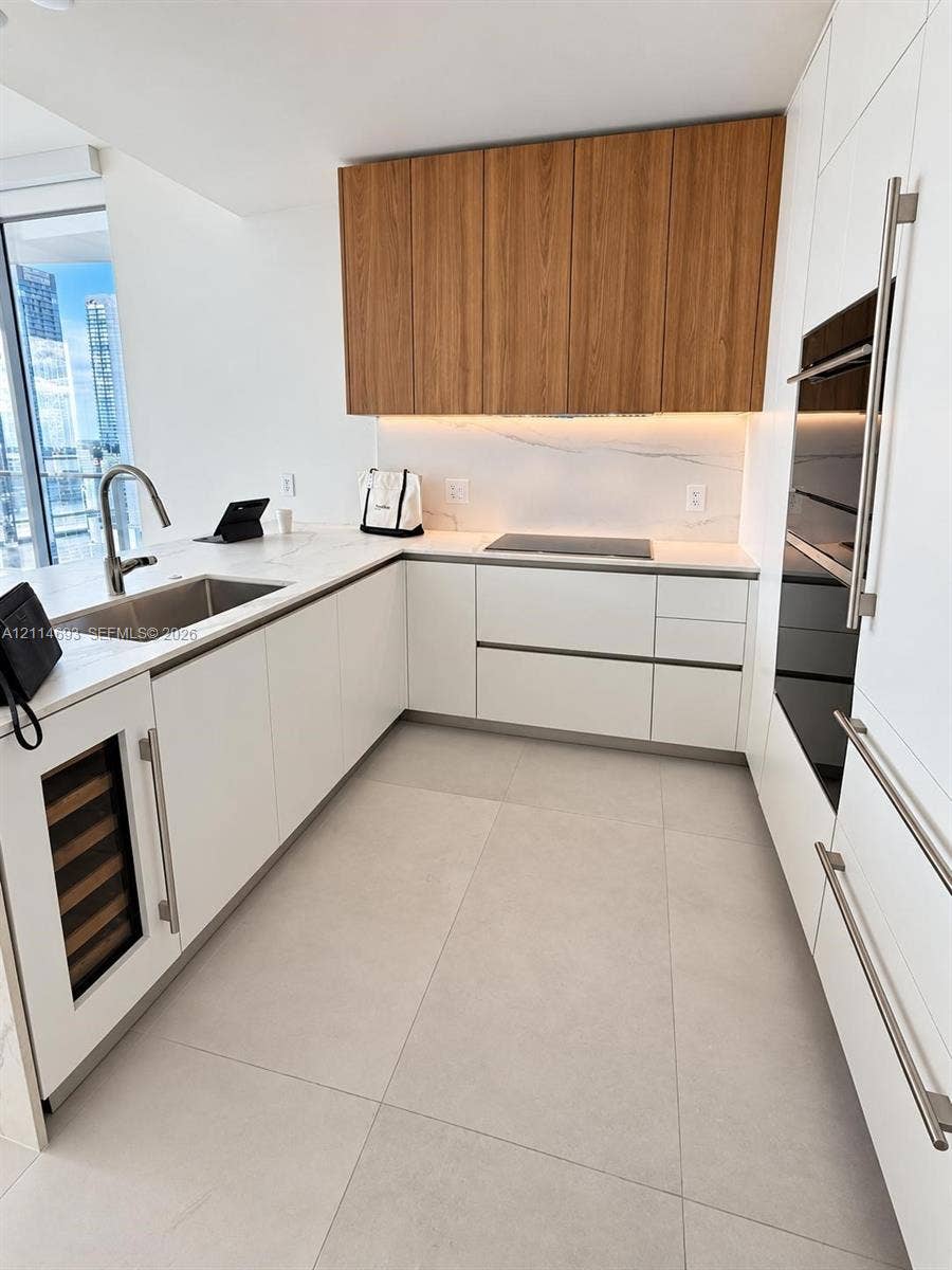 Kitchen featuring custom flat-panel cabinetry with wood-finish upper units, integrated appliances including a wine refrigerator and wall oven, stone-finish countertops, and large-format tile flooring
