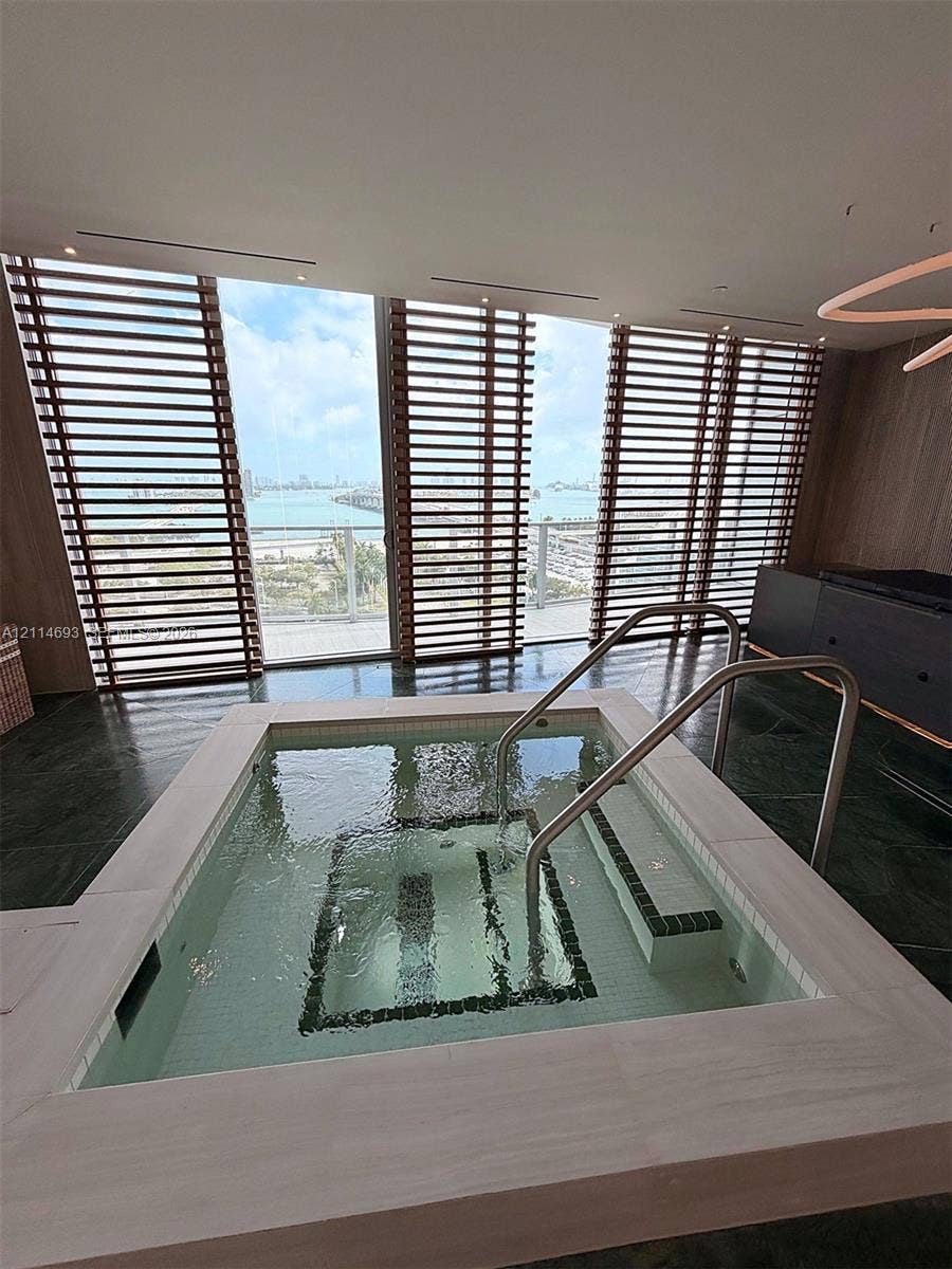 Built-in soaking tub featuring stone-tile surround, integrated steps with stainless steel handrails, floor-to-ceiling windows with wood-slat privacy screening, and dark stone-finish flooring