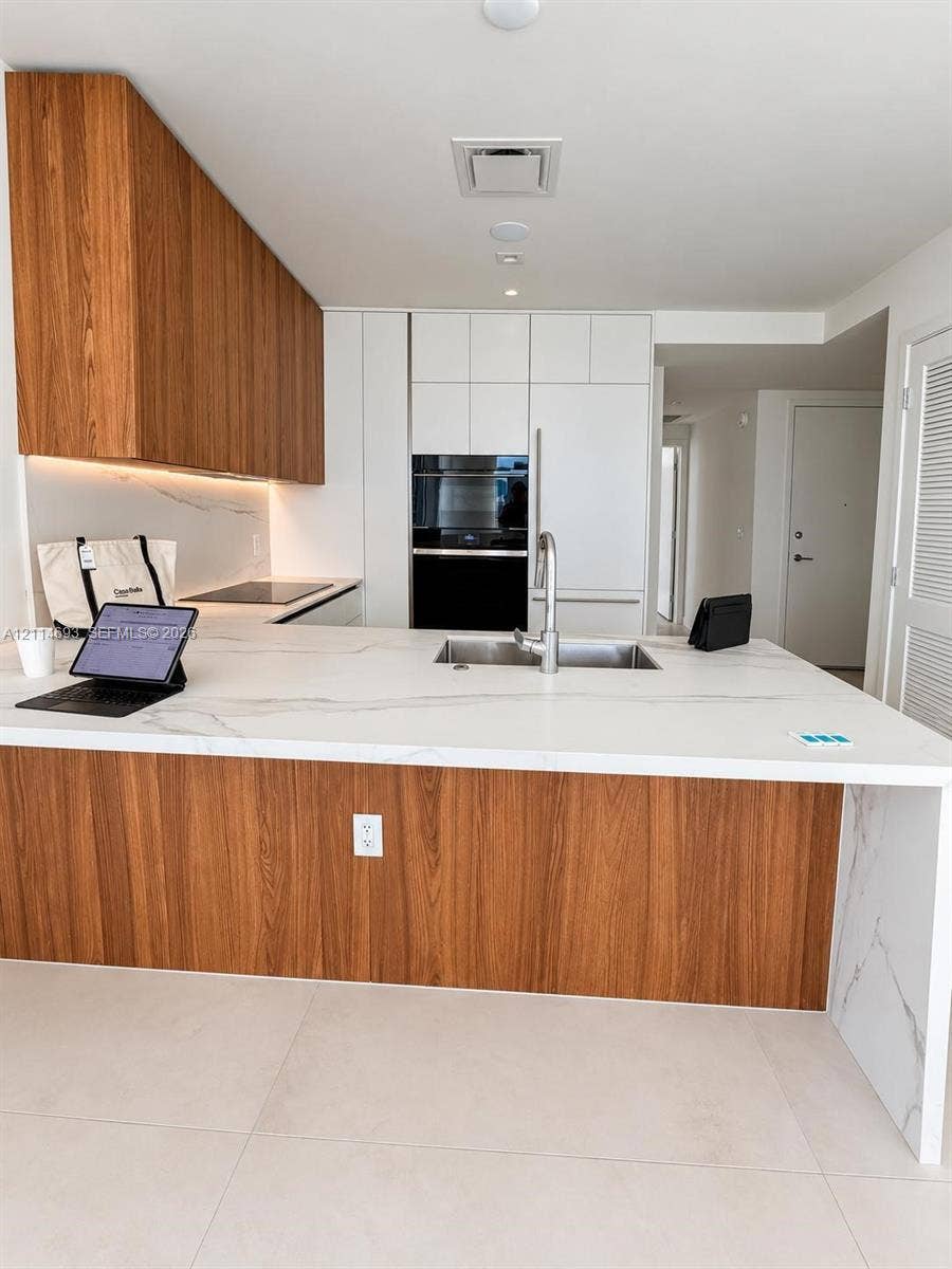 Kitchen featuring white solid-surface countertops with waterfall edge, wood-finish cabinetry, built-in wall oven, integrated cooktop, and large-format tile flooring