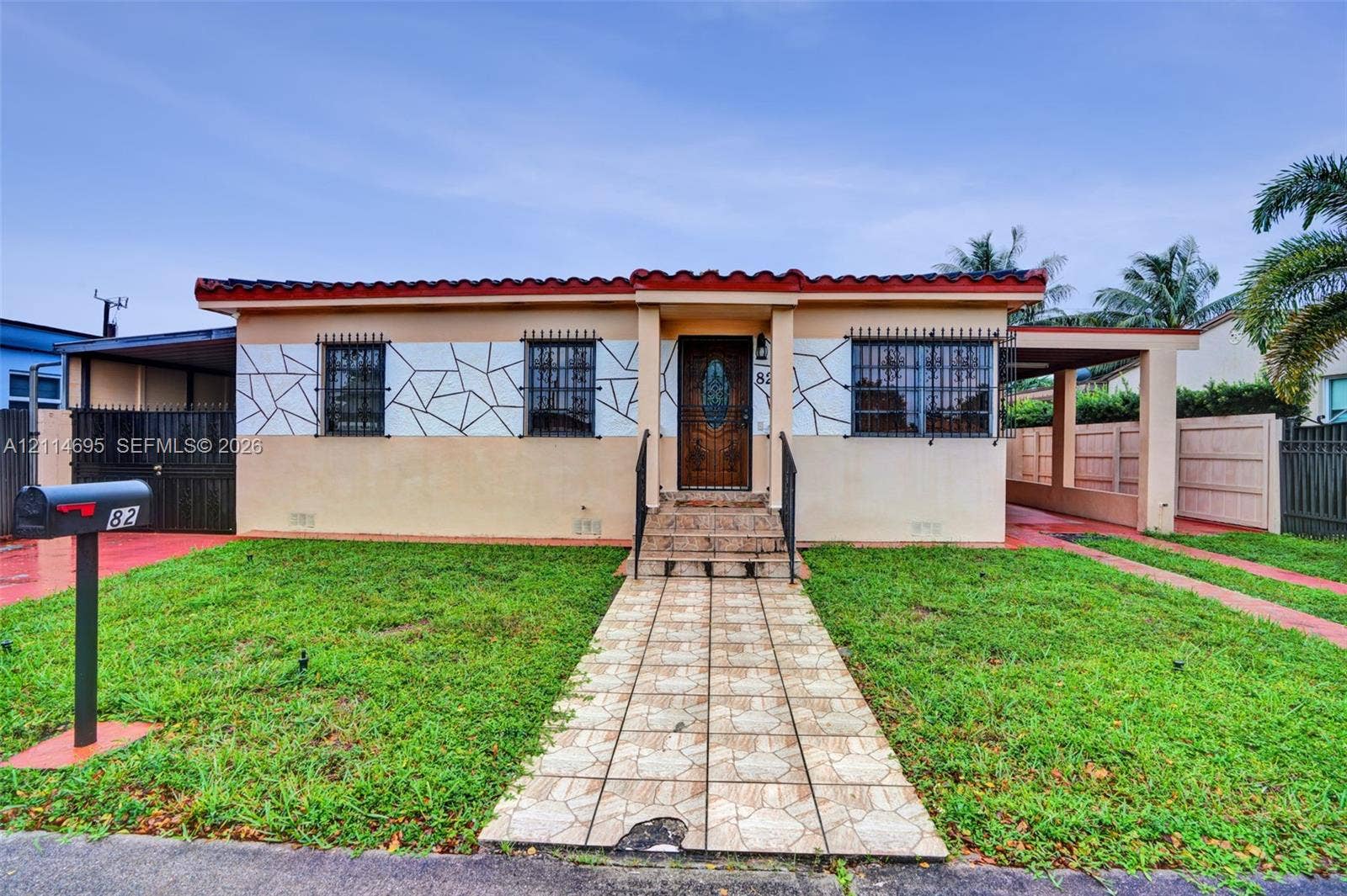 Exterior facade featuring a stucco finish with geometric decorative stonework, a low-profile red tile roof, iron security window bars, a covered carport, and a tiled walkway