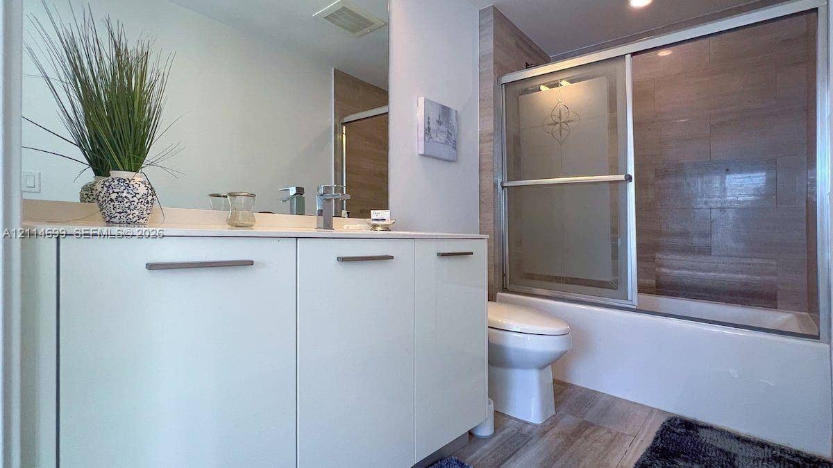 1300 Brickell Bay Drive, #3007, Miami, FL 33131 Modern vanity with white cabinetry and a solid surface countertop, featuring a single-lever faucet, large wall mirror, and a tub-shower combination with sliding glass doors and neutral-toned wall tiling