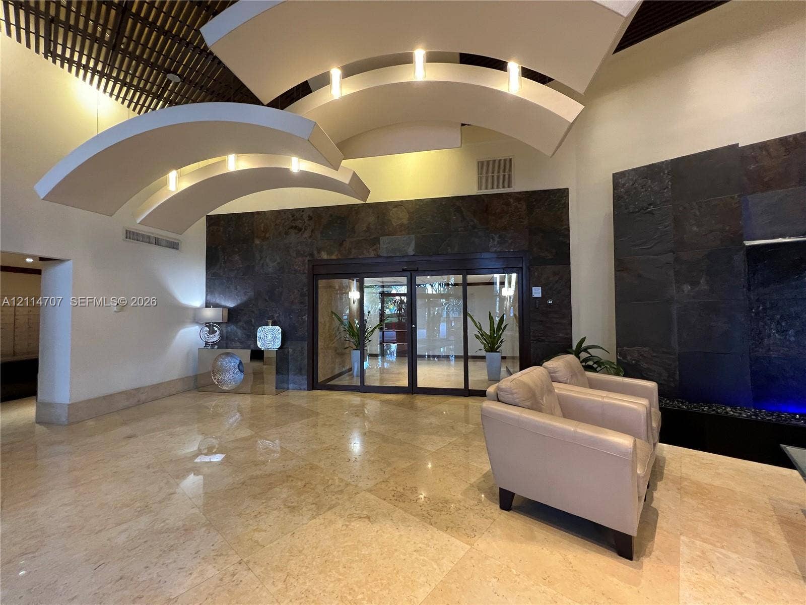 3300 NE 191st Street, #1405, Aventura, Aventura, FL 33180 Grand lobby entrance featuring tiered, curved architectural ceiling elements with recessed lighting, large-format stone floor tiles, and a prominent dark natural stone accent wall