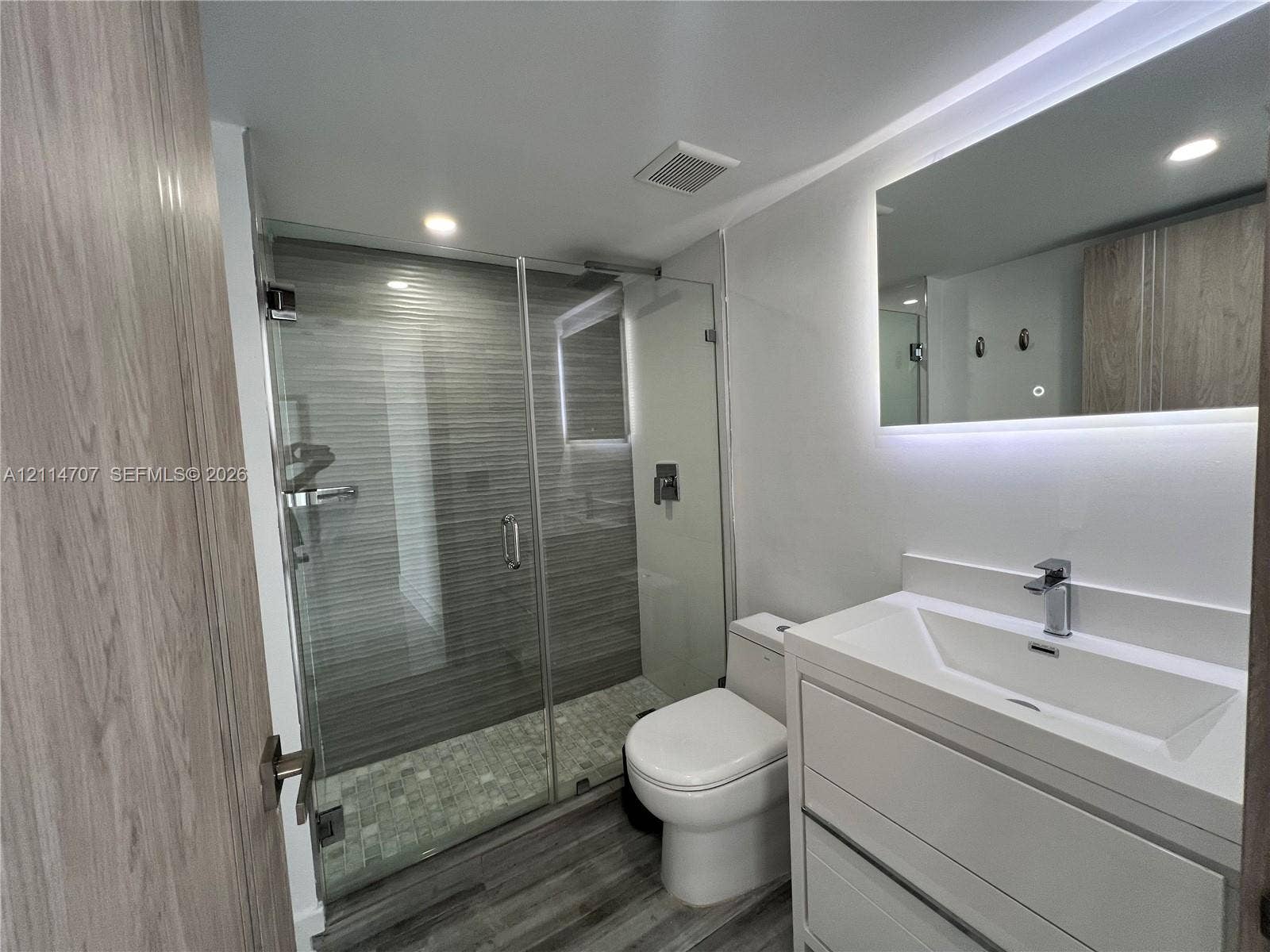 3300 NE 191st Street, #1405, Aventura, Aventura, FL 33180 Modern bathroom featuring a glass-enclosed walk-in shower with horizontal tile surround, a built-in vanity with a rectangular sink, wood-finish cabinetry, and an integrated LED-lit mirror