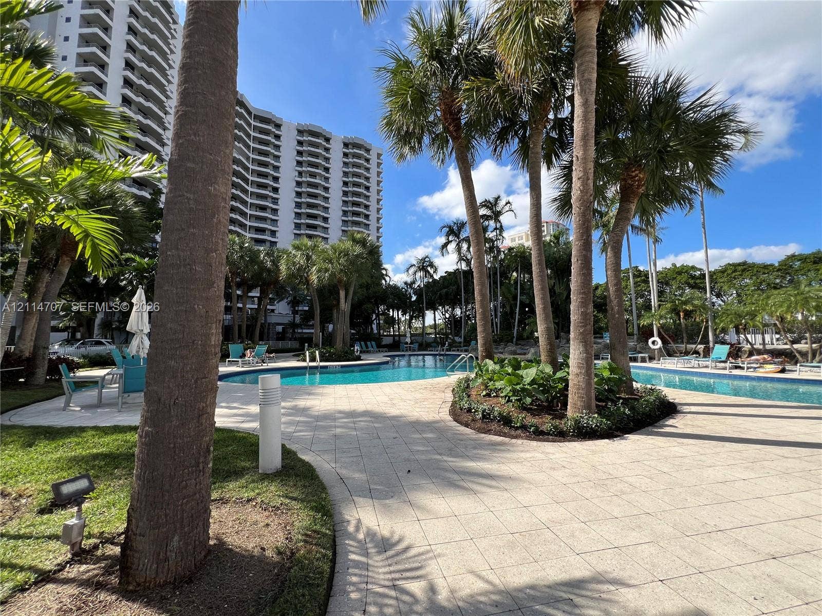 3300 NE 191st Street, #1405, Aventura, Aventura, FL 33180 Outdoor swimming pool area featuring paved stone decking, mature palm trees, landscaped garden beds, and metal ladder access