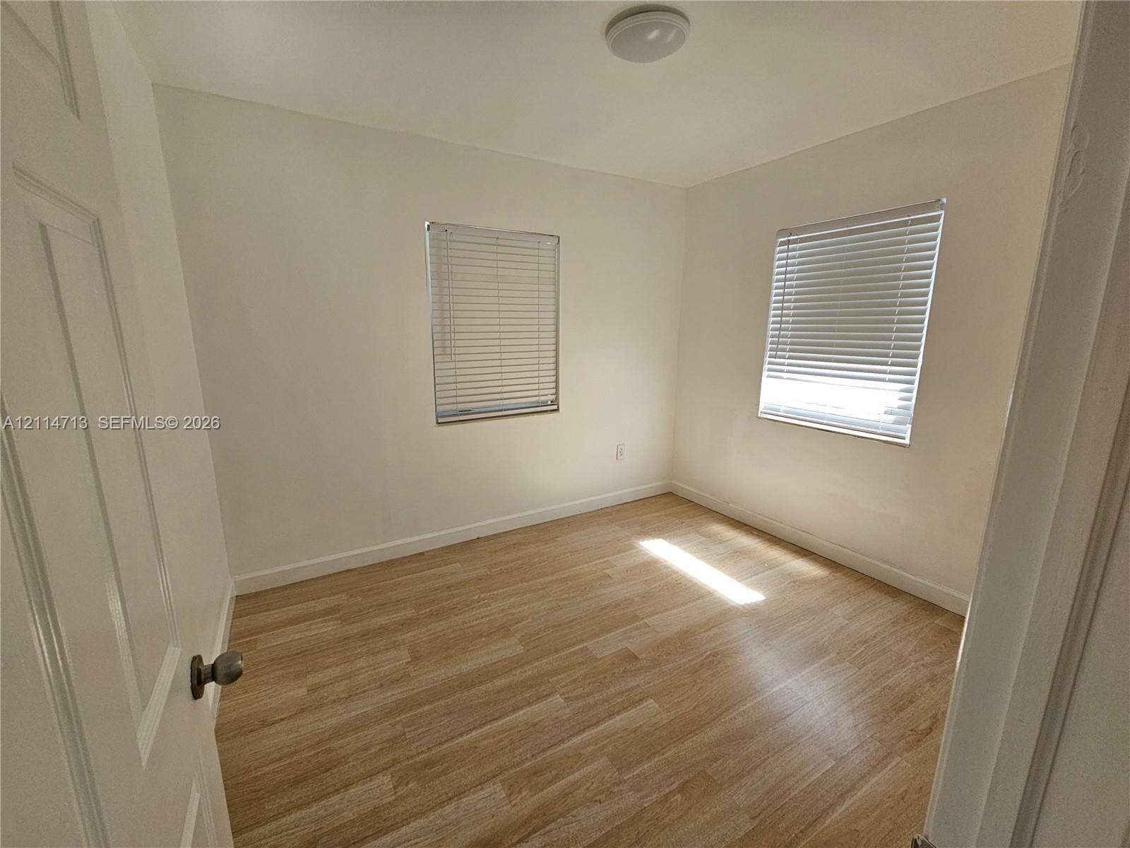 Neutral interior room featuring wood-finish flooring, two windows with horizontal blinds, and a ceiling-mounted light fixture