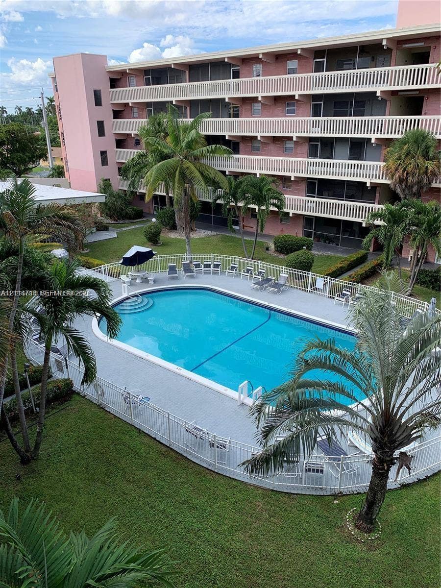 In-ground swimming pool with perimeter fencing, paved deck, manicured green space, mature palm trees, and multi-story building exterior