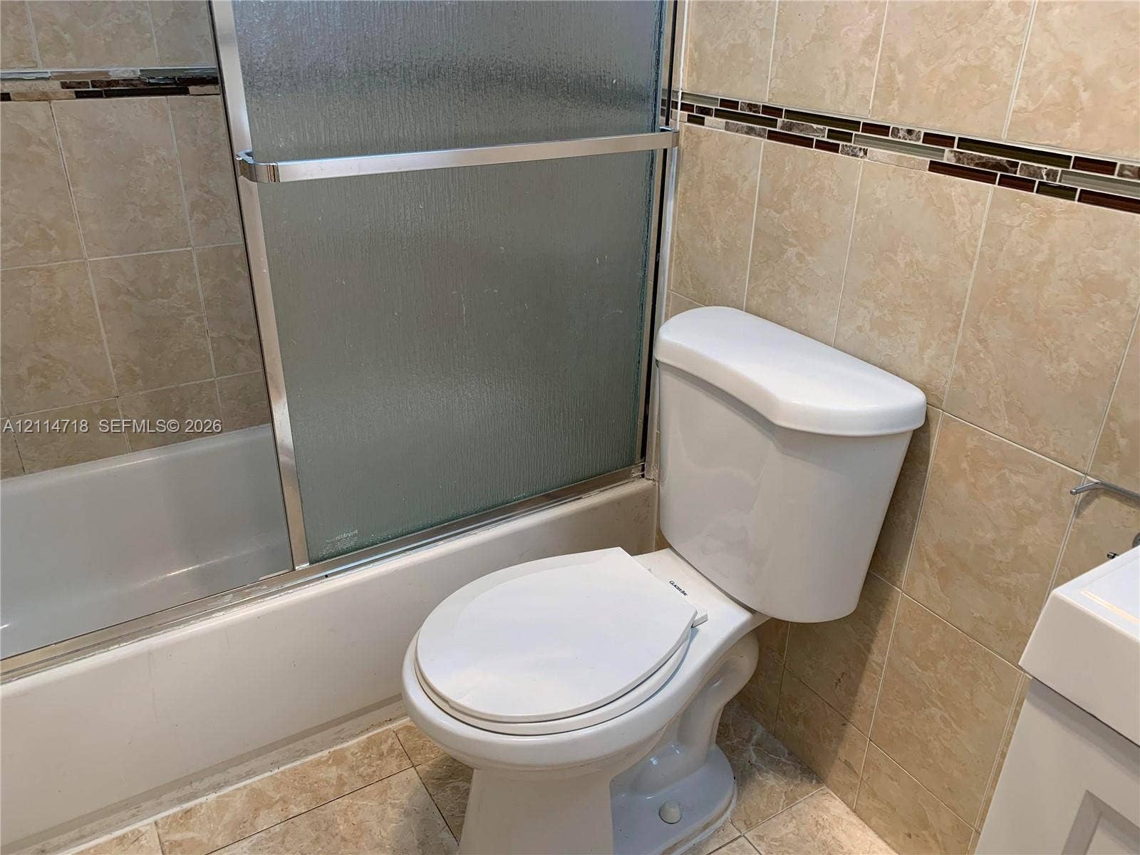Bathroom featuring floor-to-ceiling neutral ceramic tile, a glass-enclosed tub shower combination, a decorative mosaic tile accent border, and a white porcelain toilet