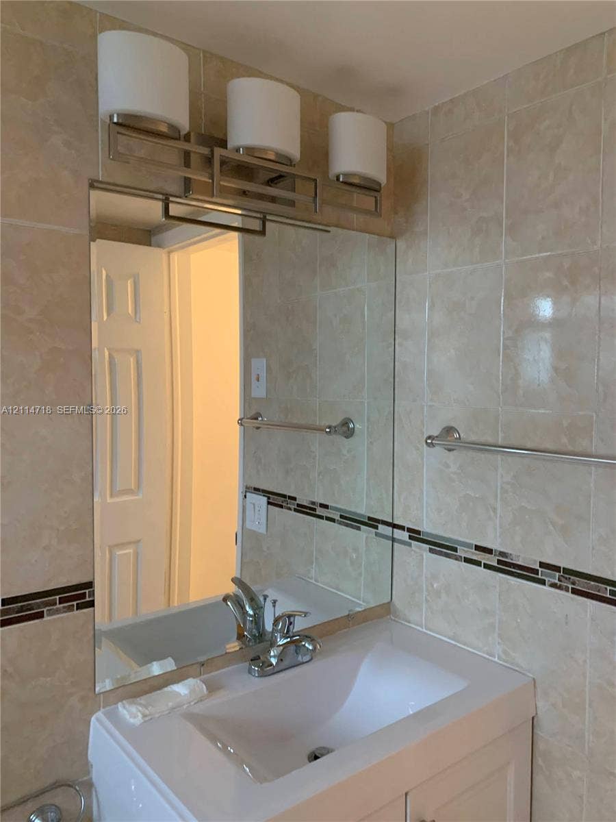 White vanity with a rectangular sink, polished chrome faucet, flat-panel mirror, three-light fixture with cylindrical glass shades, and neutral-toned wall tile with a decorative glass mosaic accent border