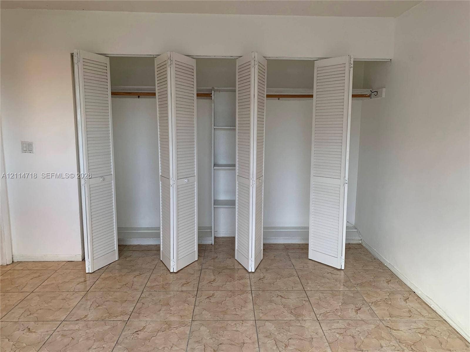 Large closet featuring white bifold louvered doors, integrated shelving, and horizontal hanging rods, complemented by neutral tile flooring