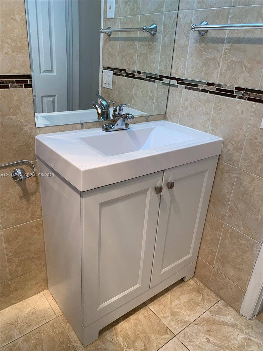 White vanity cabinet with a rectangular basin, metallic hardware, wall-mounted mirror, and tile wall surfacing featuring a decorative mosaic border