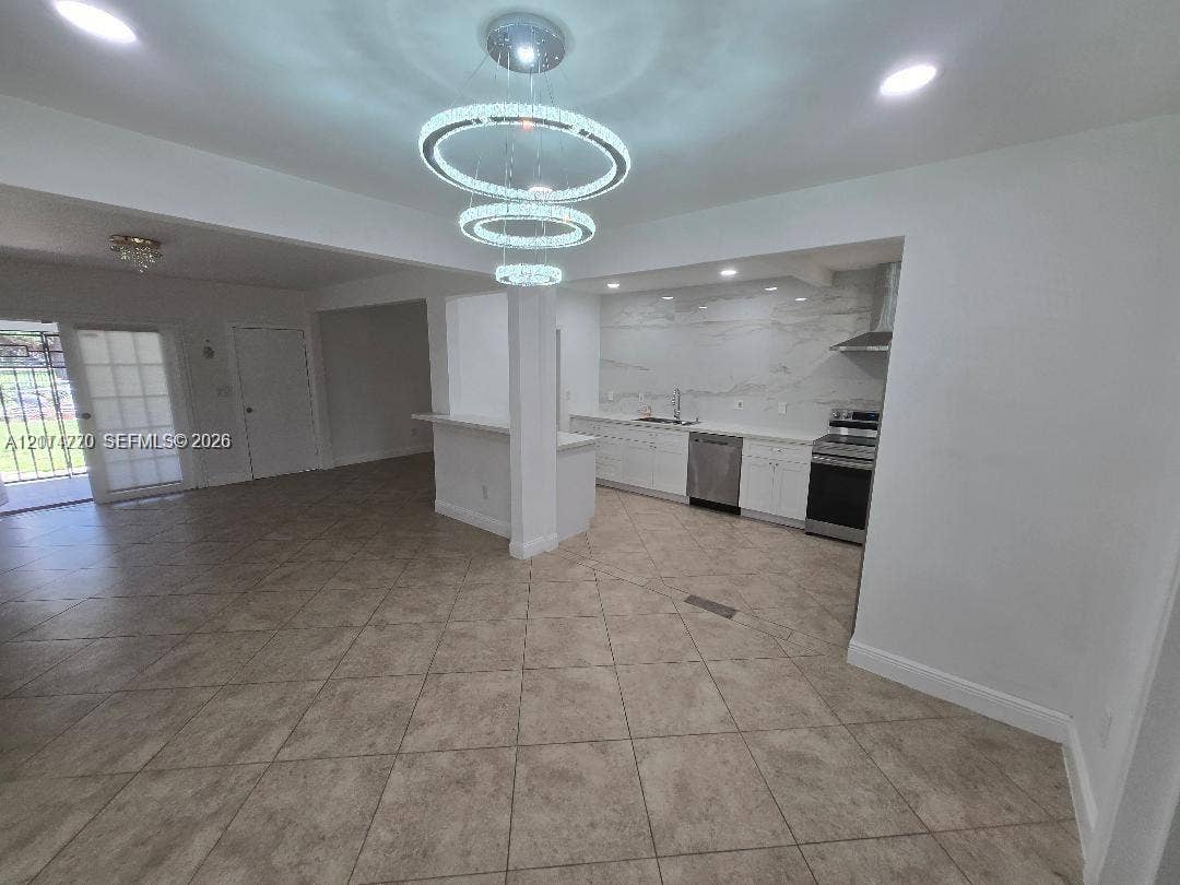 Open-concept kitchen featuring white cabinetry, stone-patterned slab backsplash, stainless steel appliances, tiered crystal chandelier, and tile flooring throughout