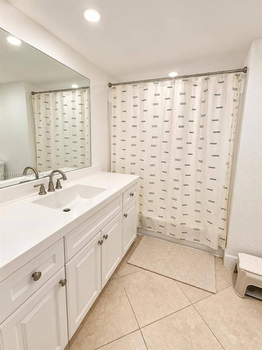 White cabinetry vanity featuring a rectangular integrated sink, white countertop, brushed metal faucet, and full-width wall mirror, complemented by tile flooring