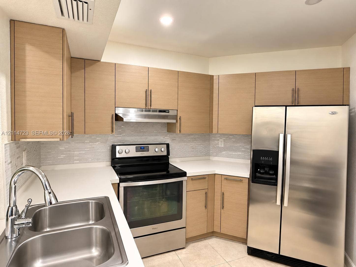Kitchen featuring flat-panel wood-finish cabinetry, solid surface countertops, a mosaic tile backsplash, stainless steel appliances, and a dual-basin stainless steel sink with a gooseneck faucet