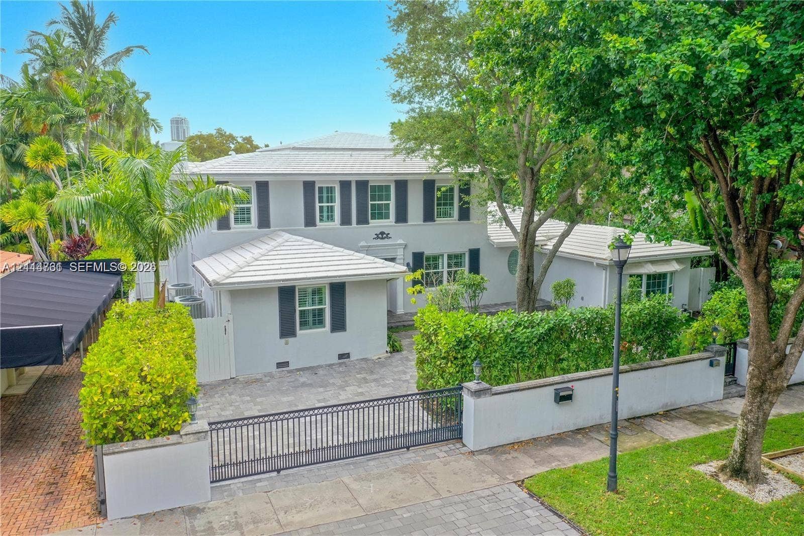 Two-story exterior featuring a light-toned stucco faade, metal roof, shuttered windows, paver driveway, and a gated perimeter wall