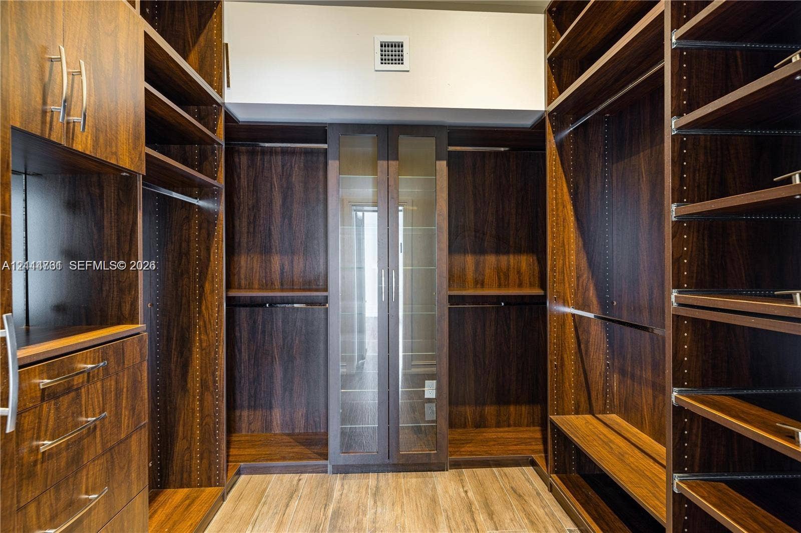 Custom walk-in closet featuring wood-finish cabinetry, built-in drawer units, adjustable shelving, integrated hanging rods, and glass-front cabinetry doors with metal hardware