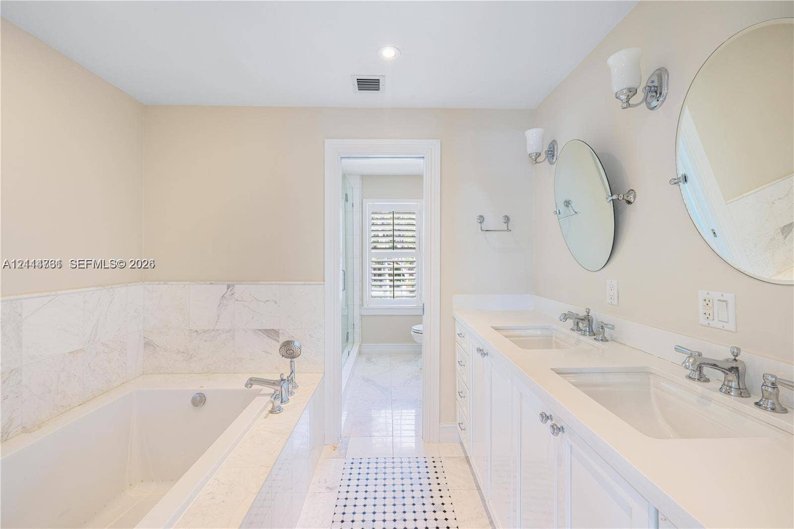 Double vanity with integrated sinks, white cabinetry, polished metal fixtures, and wall-mounted mirrors, featuring a soaking tub with stone tile surround and stone flooring