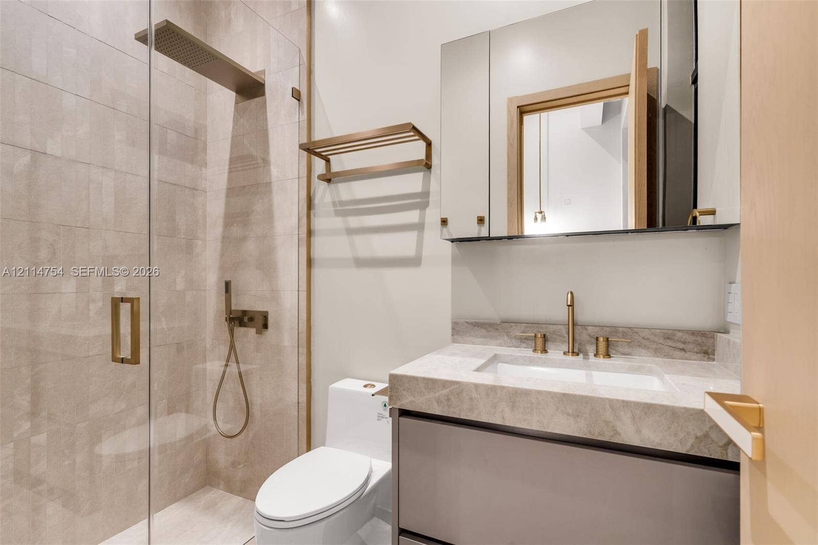 6731 SW 64th Avenue, South Miami, FL 33143 Modern vanity with a natural stone countertop and brass-finish faucet, featuring a mirrored medicine cabinet, a wall-mounted brass towel rack, and a glass-enclosed shower with decorative hexagonal tile wall covering and a rainfall showerhead