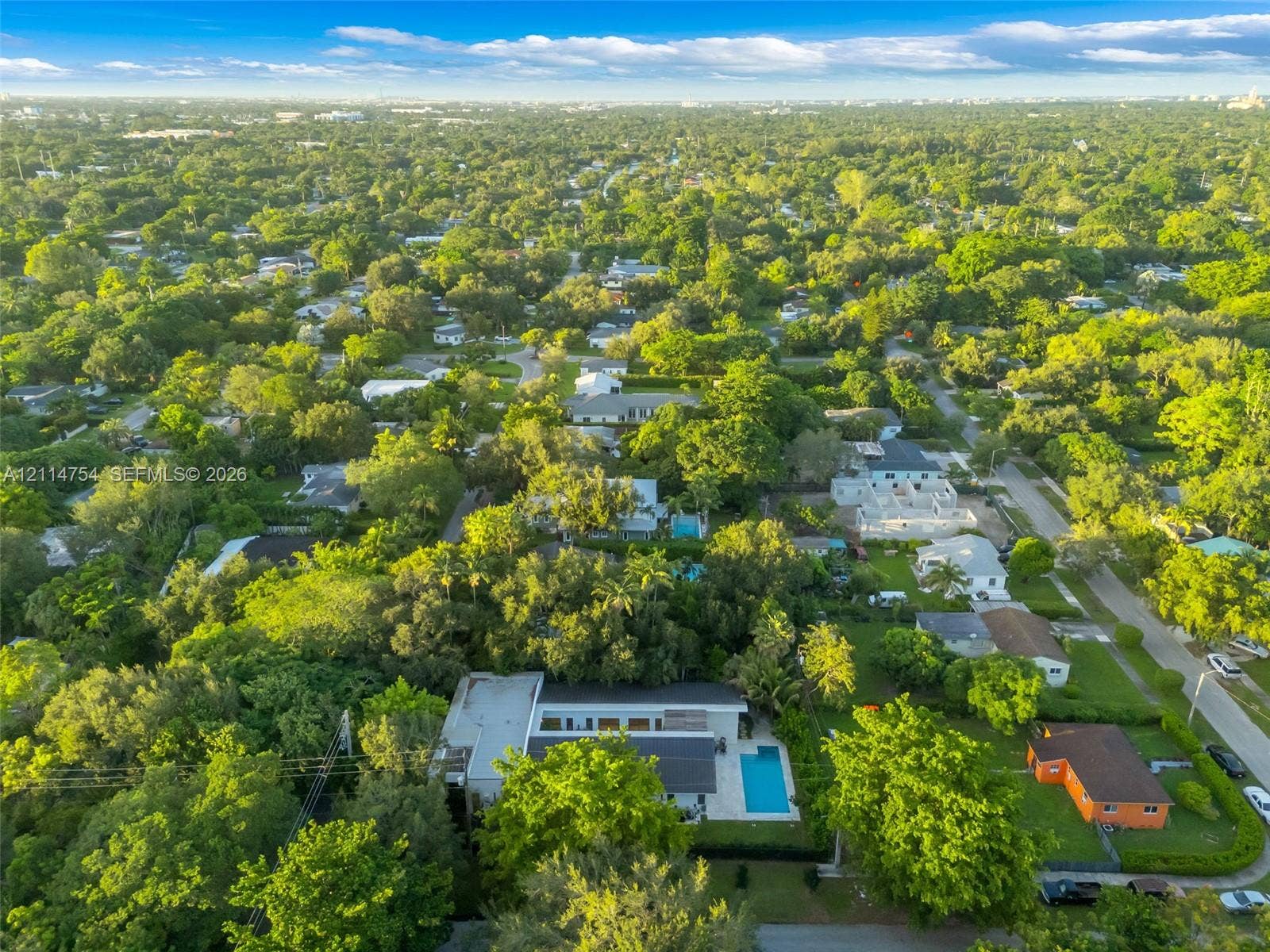 6731 SW 64th Avenue, South Miami, FL 33143 Aerial perspective featuring a flat-roof residence, a rectangular swimming pool, and mature canopy trees, set against a distant cityscape backdrop