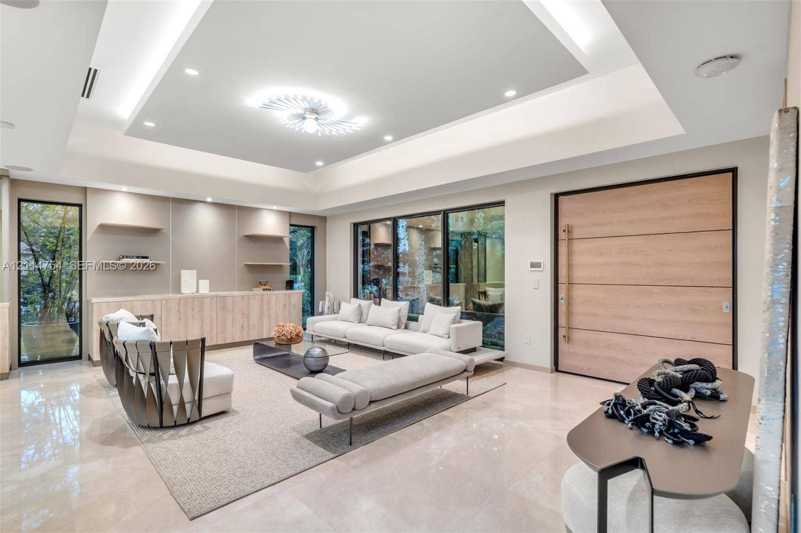 6731 SW 64th Avenue, South Miami, FL 33143 Expansive living area featuring a tray ceiling with recessed lighting, large-format polished flooring, floor-to-ceiling glass wall panels with dark frames, and distinctive modern tiered ceiling-mounted lighting