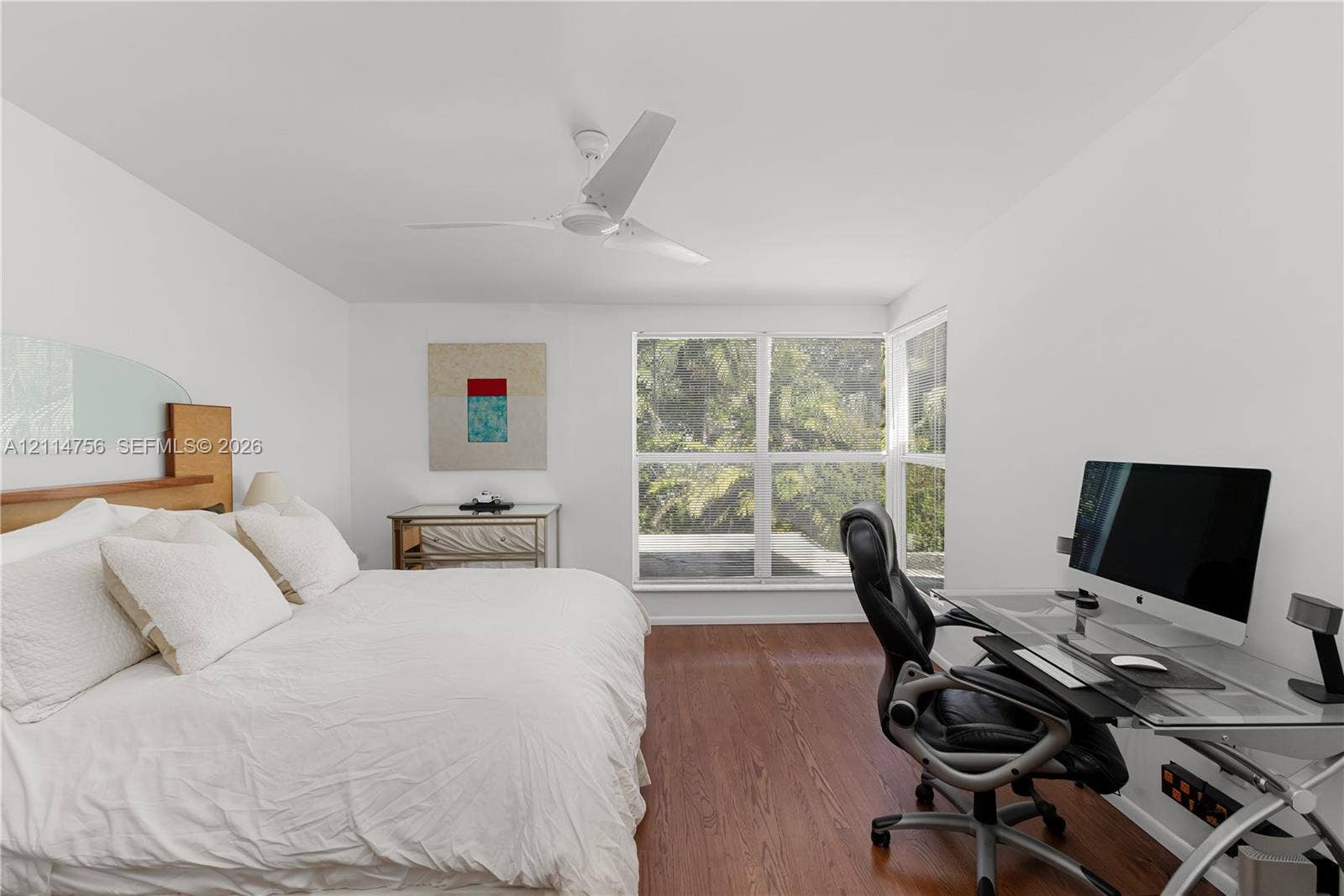 Room featuring wood-finish flooring, white walls, a ceiling fan, and large windows with exterior greenery