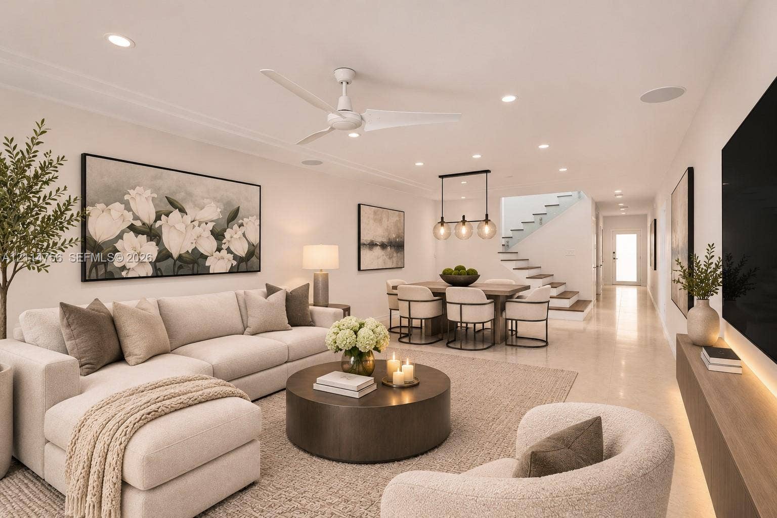 Open-concept interior featuring light-toned flooring, recessed ceiling lighting, a contemporary three-blade ceiling fan, a linear wood-finish wall-mounted media console with integrated lighting, and a staircase with glass railings