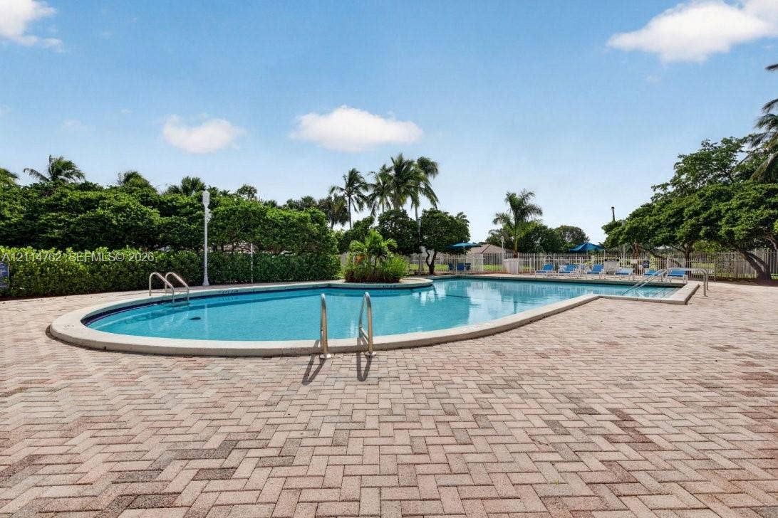 662 NW 170th Terrace, Pembroke Pines, FL 33028 In-ground swimming pool featuring a curved design, metal handrails, and a surrounding herringbone-patterned paver deck bordered by established hedging and mature palms