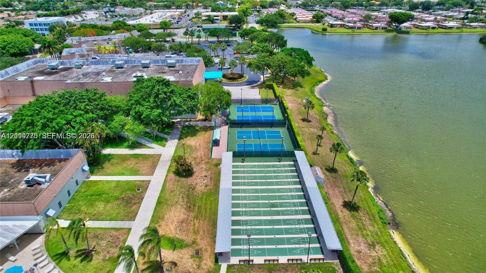 653 N Saxony N, #653, Delray Beach, FL 33446 Aerial perspective of recreational facilities including two fenced tennis courts and multiple shuffleboard courts situated along a water feature with mature landscaping and paved pedestrian walkways