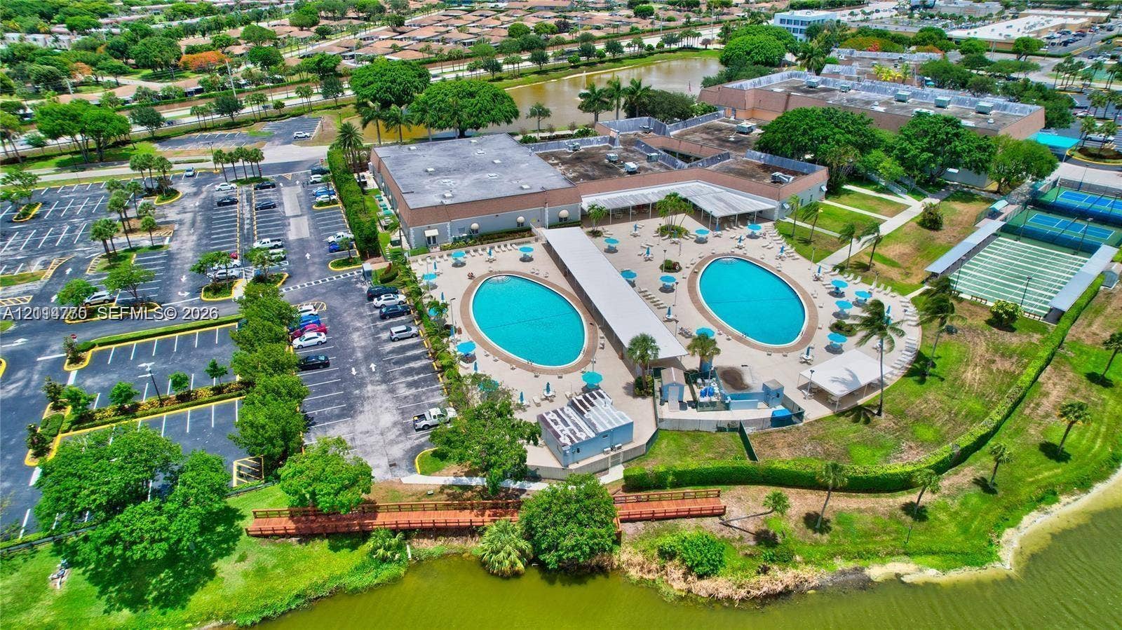 653 N Saxony N, #653, Delray Beach, FL 33446 Aerial view of recreational facility featuring two outdoor swimming pools, multi-use tennis courts, paved parking area, and waterfront access with a wooden boardwalk