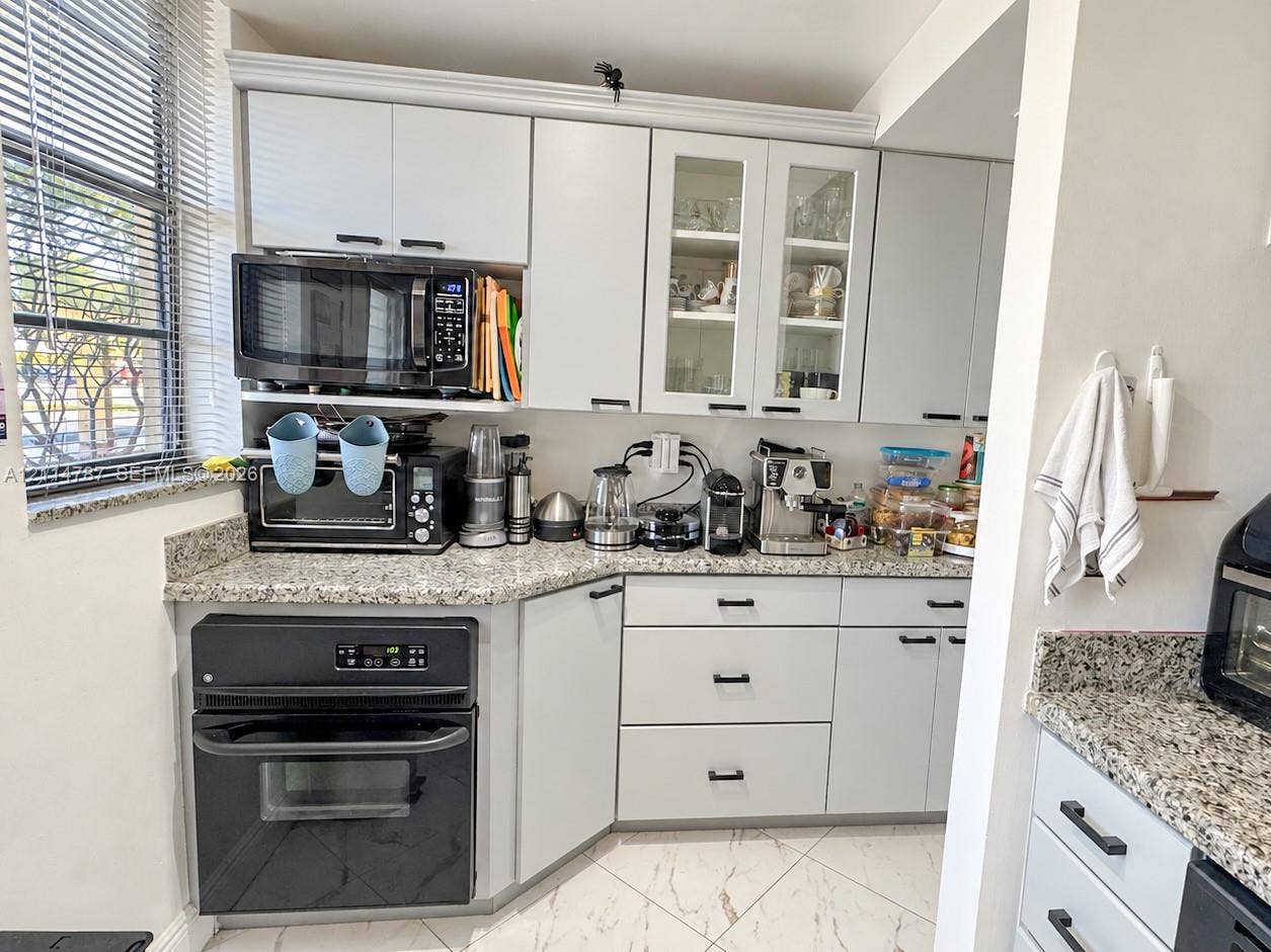 Light gray shaker-style cabinetry with glass-paneled uppers, stone-patterned countertops, integrated wall oven, and tile flooring