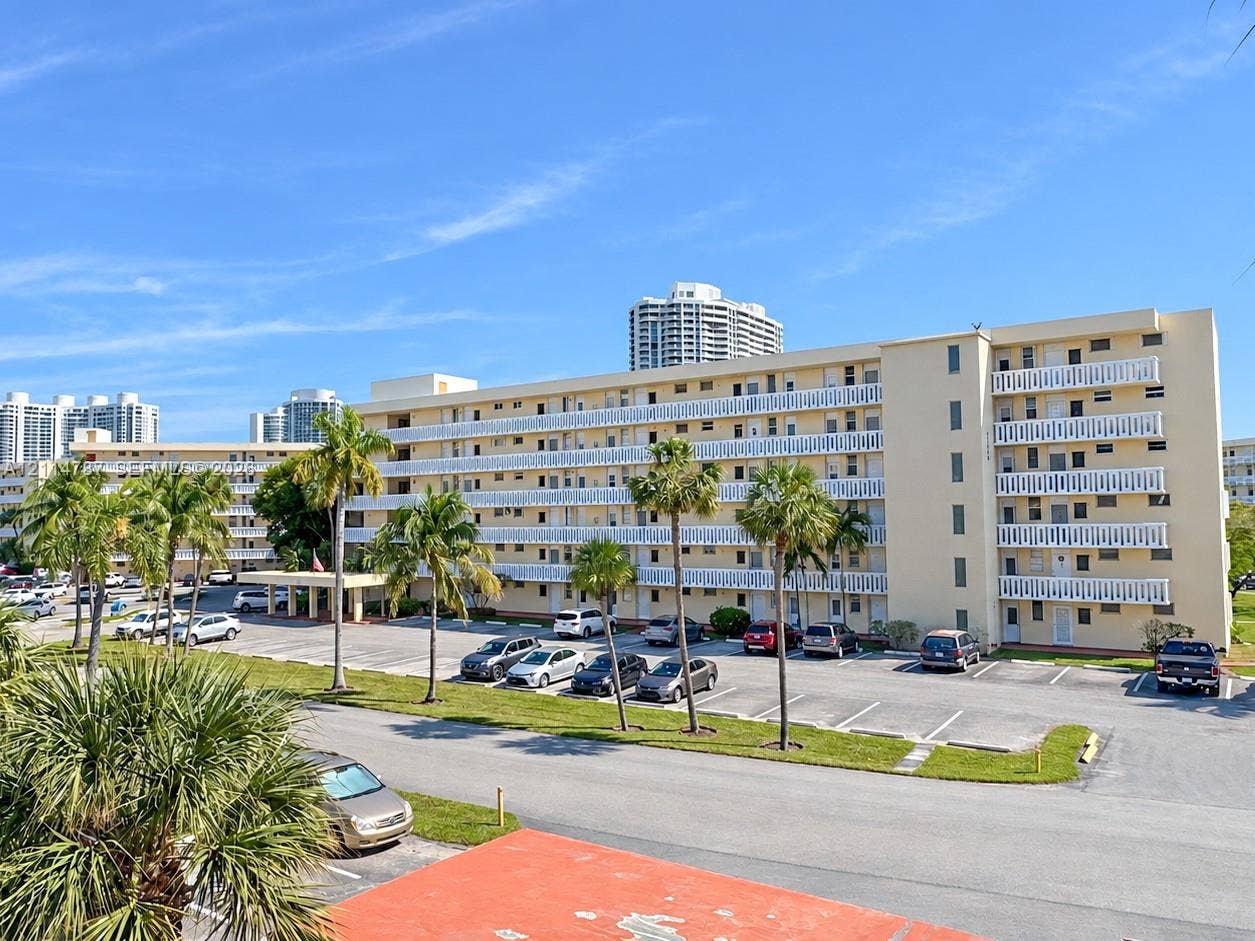 Multi-story residential building featuring exterior balconies with decorative lattice railings, ground-level surface parking, and mature palm trees