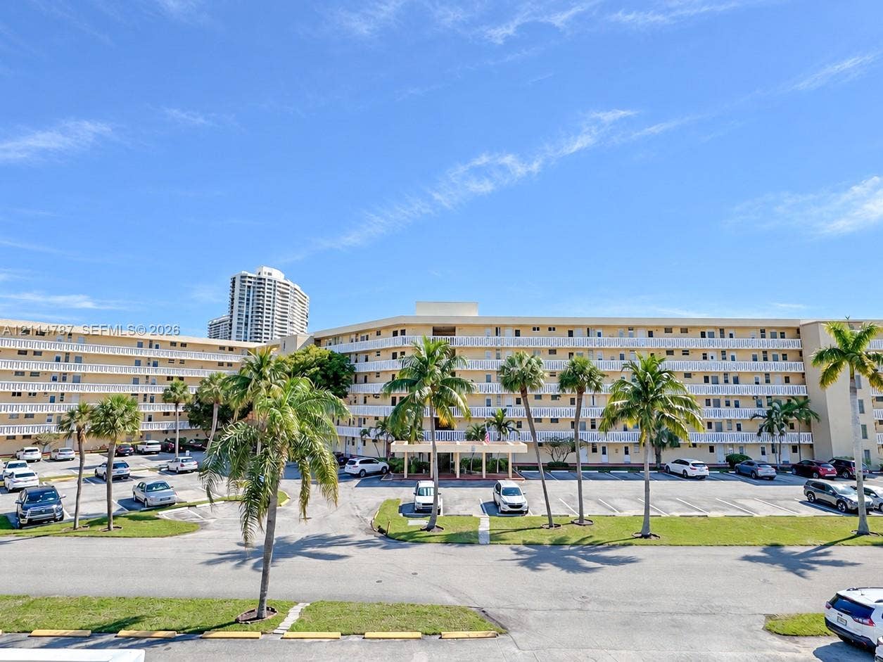 Multi-story residential complex featuring continuous exterior balcony railings, a central covered porte-cochre entry, surface parking, and mature palm tree landscaping