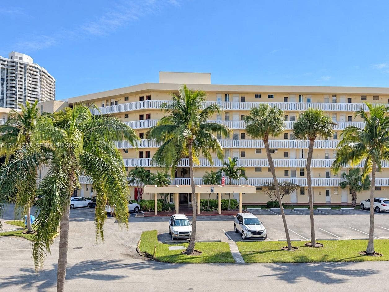 Mid-rise building exterior featuring stucco cladding, open-air balcony railings, a covered entrance, and landscaped grounds with mature palm trees