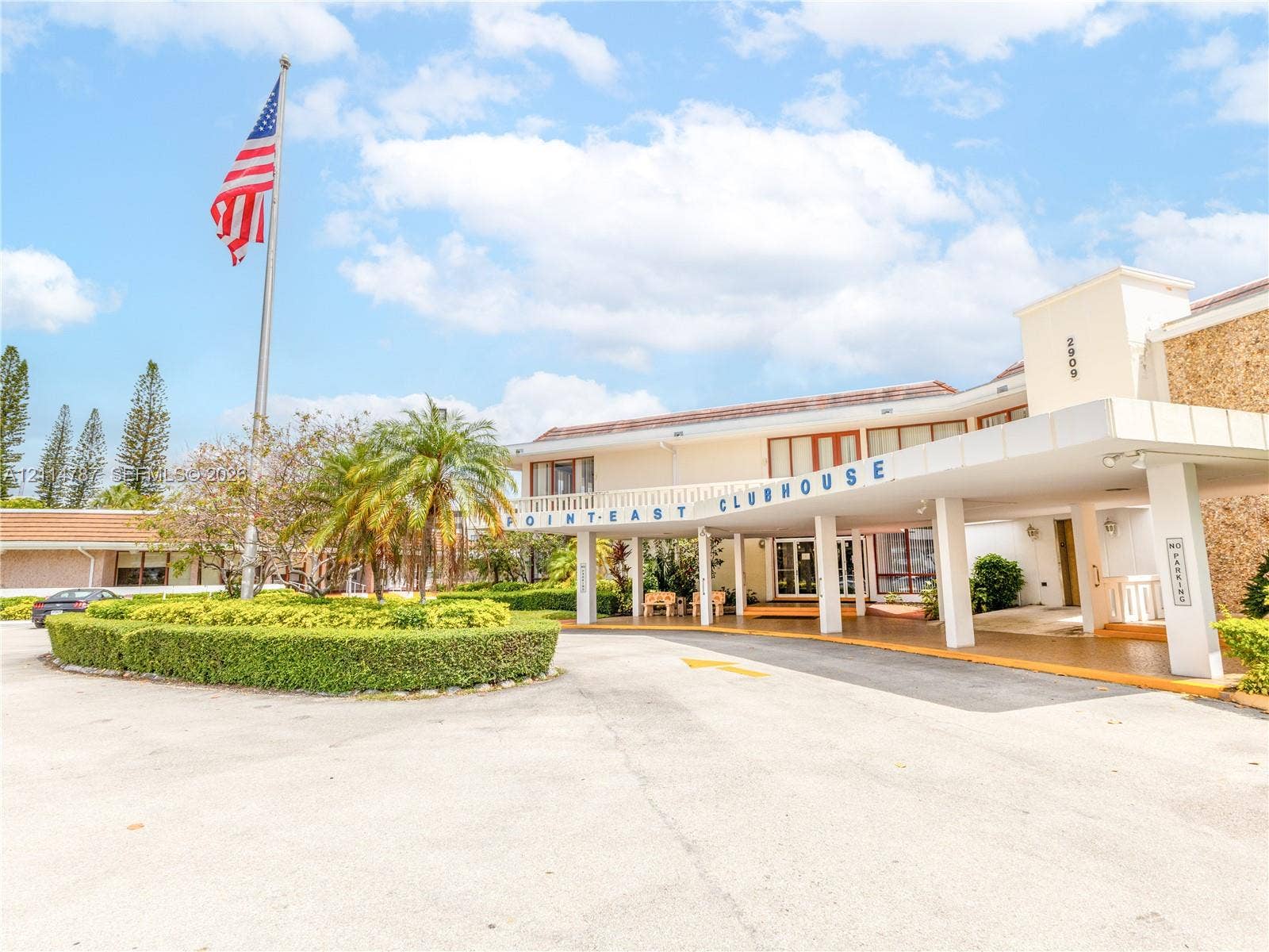 Clubhouse exterior featuring a covered porte-cochre, stone masonry facade, manicured landscaping, and asphalt parking area