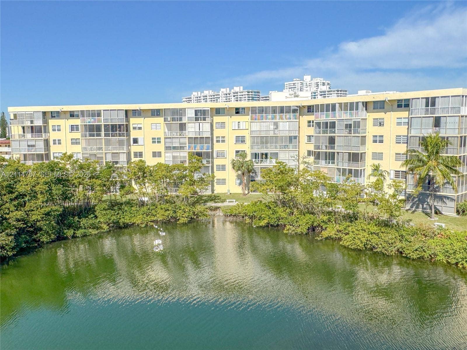 Multi-story building featuring screened balconies, light-colored exterior facade, mature landscaping, and waterfront positioning