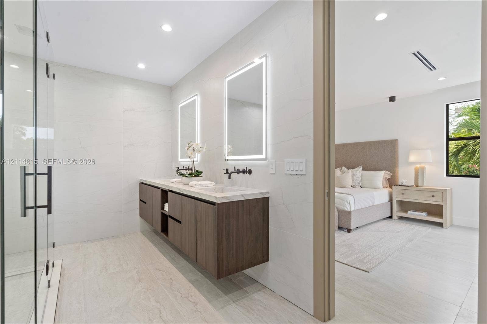 Floating wood-finish vanity with dual sinks, stone-surface countertop, matte black wall-mounted faucets, large illuminated mirrors, and glass-enclosed shower with large-format tile wall cladding