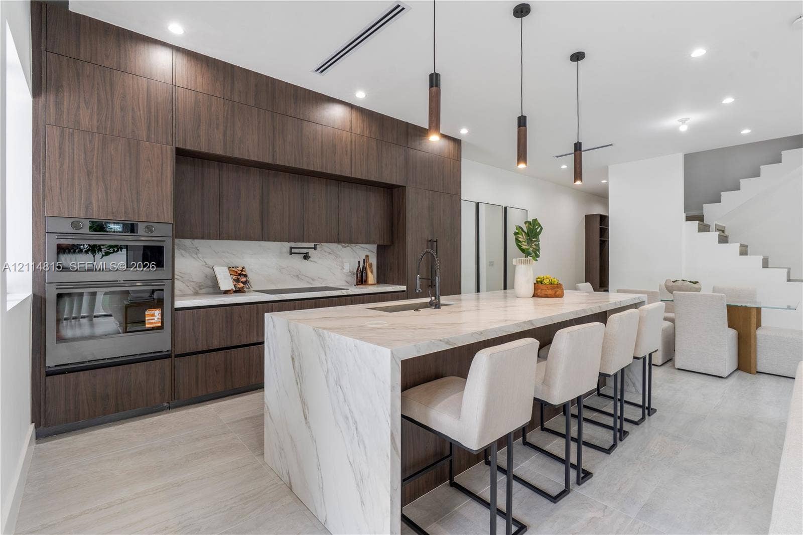 Kitchen featuring dark wood-finish cabinetry, a waterfall-edge stone island with an integrated sink, built-in double wall ovens, a pot filler faucet, and pendant lighting