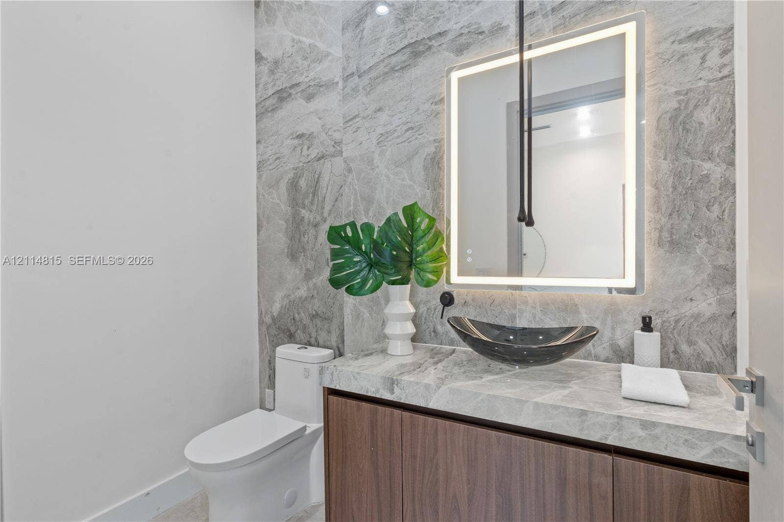 Modern vanity with wood-finish cabinetry, stone countertop, vessel sink, and wall-mounted faucet, complemented by full-height stone wall tiling and a rectangular backlit LED mirror