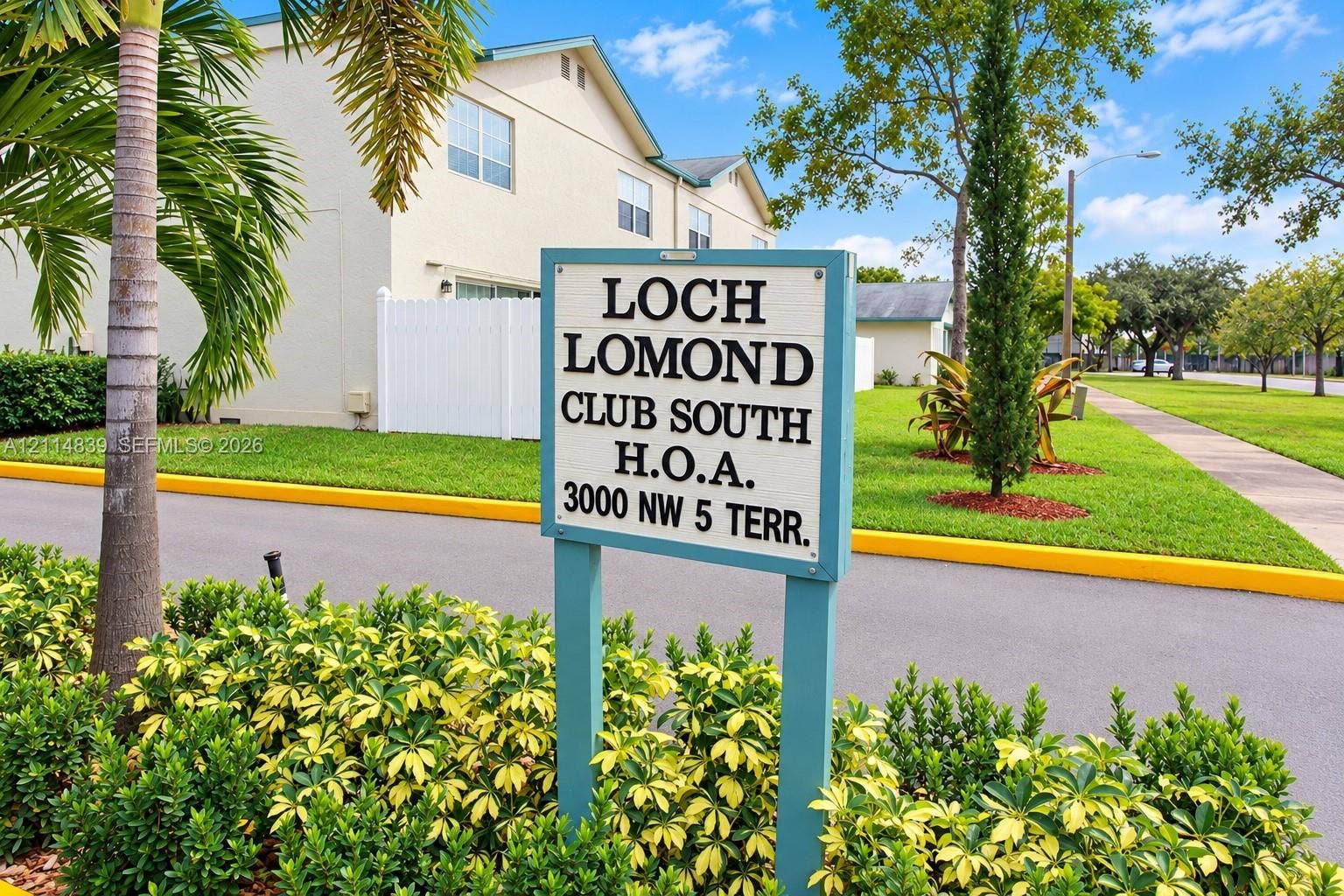 3000 NW 5th Terrace, #131, Pompano Beach, Pompano Beach, FL 33064 Exterior featuring a light-toned stucco facade, white vinyl fencing, paved asphalt pathways, and mature palm and cypress landscaping