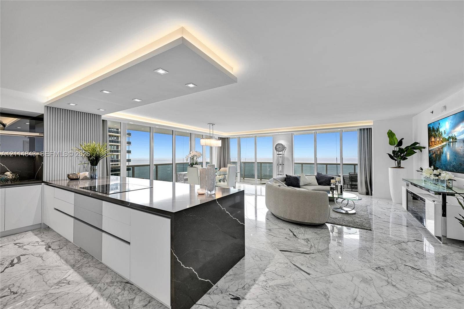 Open-concept living space featuring stone flooring, floor-to-ceiling windows, a center kitchen island with a dark stone waterfall countertop, recessed ceiling lighting, and custom cabinetry
