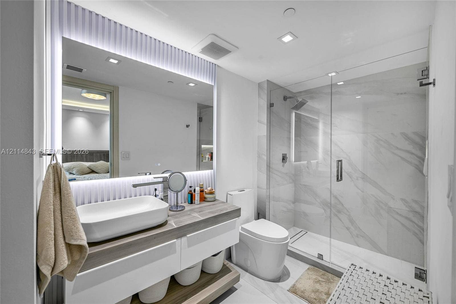 Floating vanity with a rectangular vessel sink and wood-finish countertop, full-height backlit mirror, walk-in glass shower enclosure with stone-finish tile surround, and recessed ceiling lighting