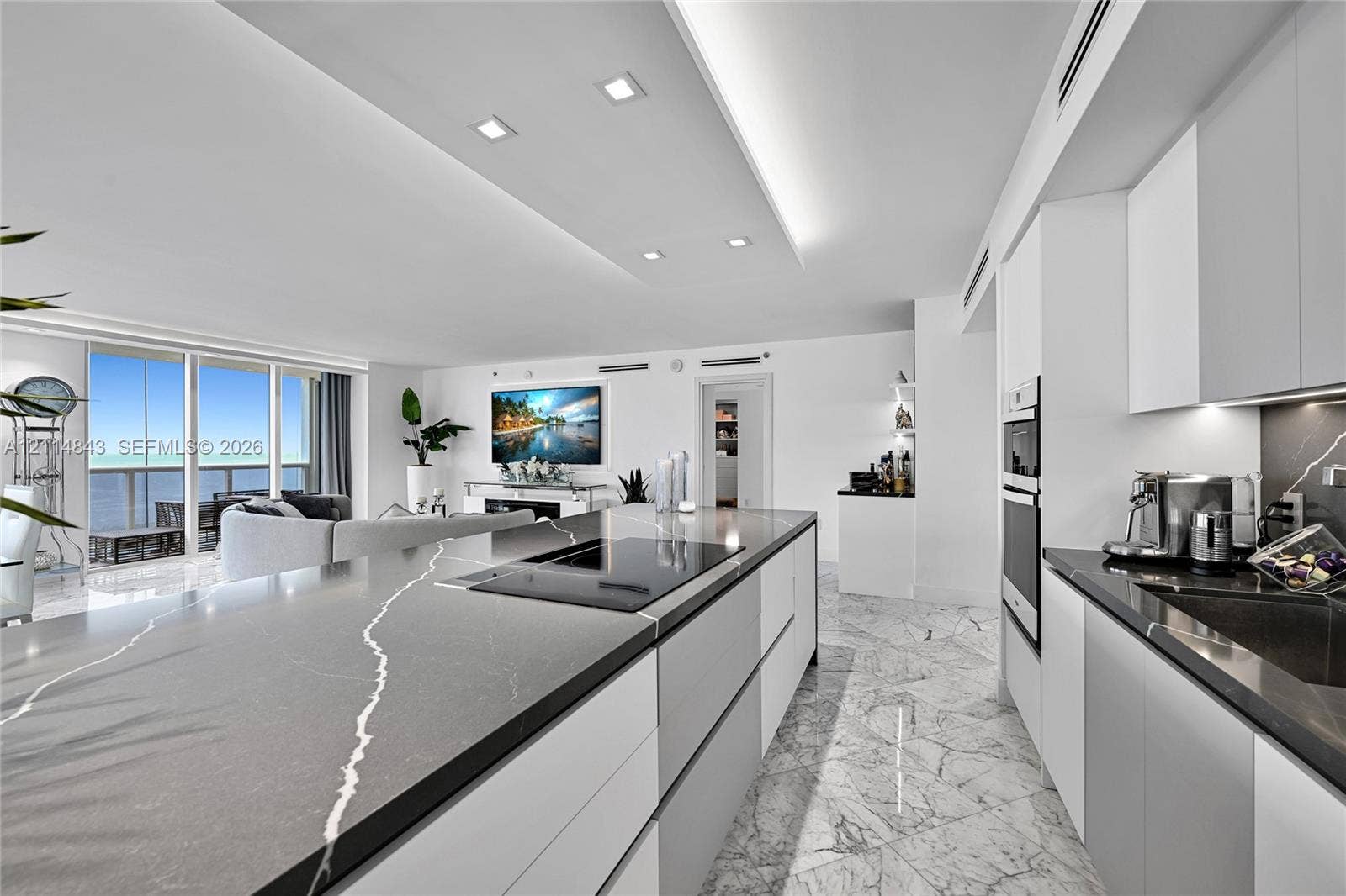 Modern kitchen featuring a large central island with veined stone countertop and integrated cooktop, handle-less cabinetry, built-in double ovens, and polished stone-finish flooring