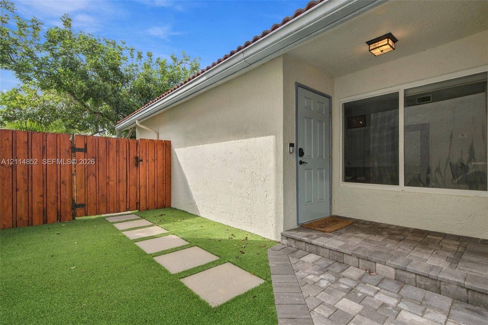 10780 NW 10th Street, Plantation, FL 33322 Exterior entryway featuring a stucco facade, a stone-paver porch, a wood-finish fence, and a stepped paver walkway set into synthetic turf