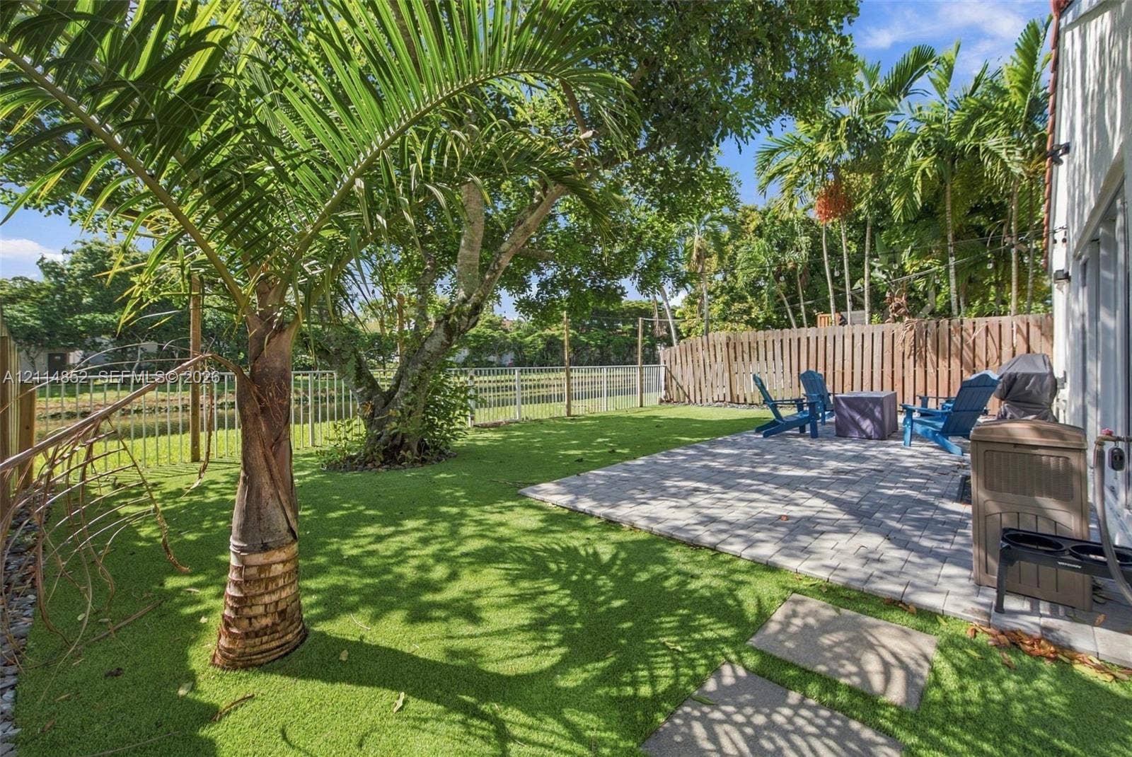 10780 NW 10th Street, Plantation, FL 33322 Private backyard featuring a paved stone patio, synthetic turf lawn, mature palm trees, and perimeter wood fencing