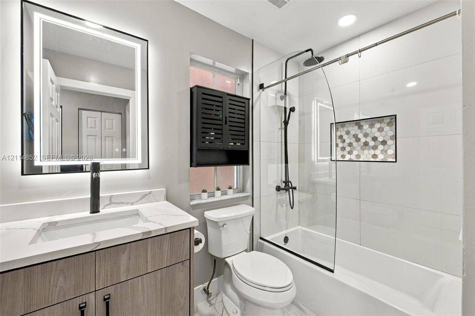 10780 NW 10th Street, Plantation, FL 33322 Bathroom featuring a wood-finish vanity with a stone-top integrated sink, matte black faucet, LED-backlit mirror, and a tub-shower combination with a glass panel and hexagon tile recessed niche