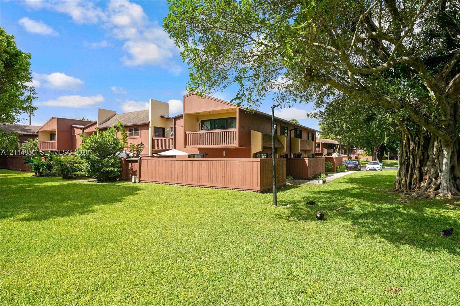 Miami, FL 33173 Multi-level residential exterior featuring horizontal wood siding, private balconies with wood railings, enclosed courtyard fencing, and expansive green space with mature trees