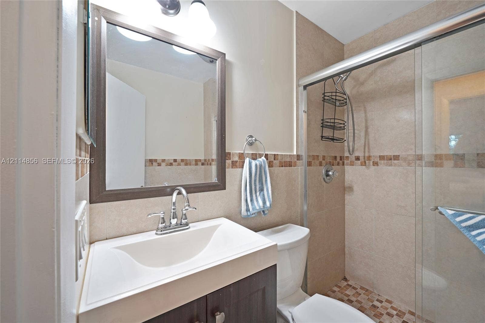 Miami, FL 33173 Bathroom featuring a dark wood-finish vanity with an integrated white sink, framed wall mirror, tiled shower with a sliding glass door, and decorative mosaic tile accents