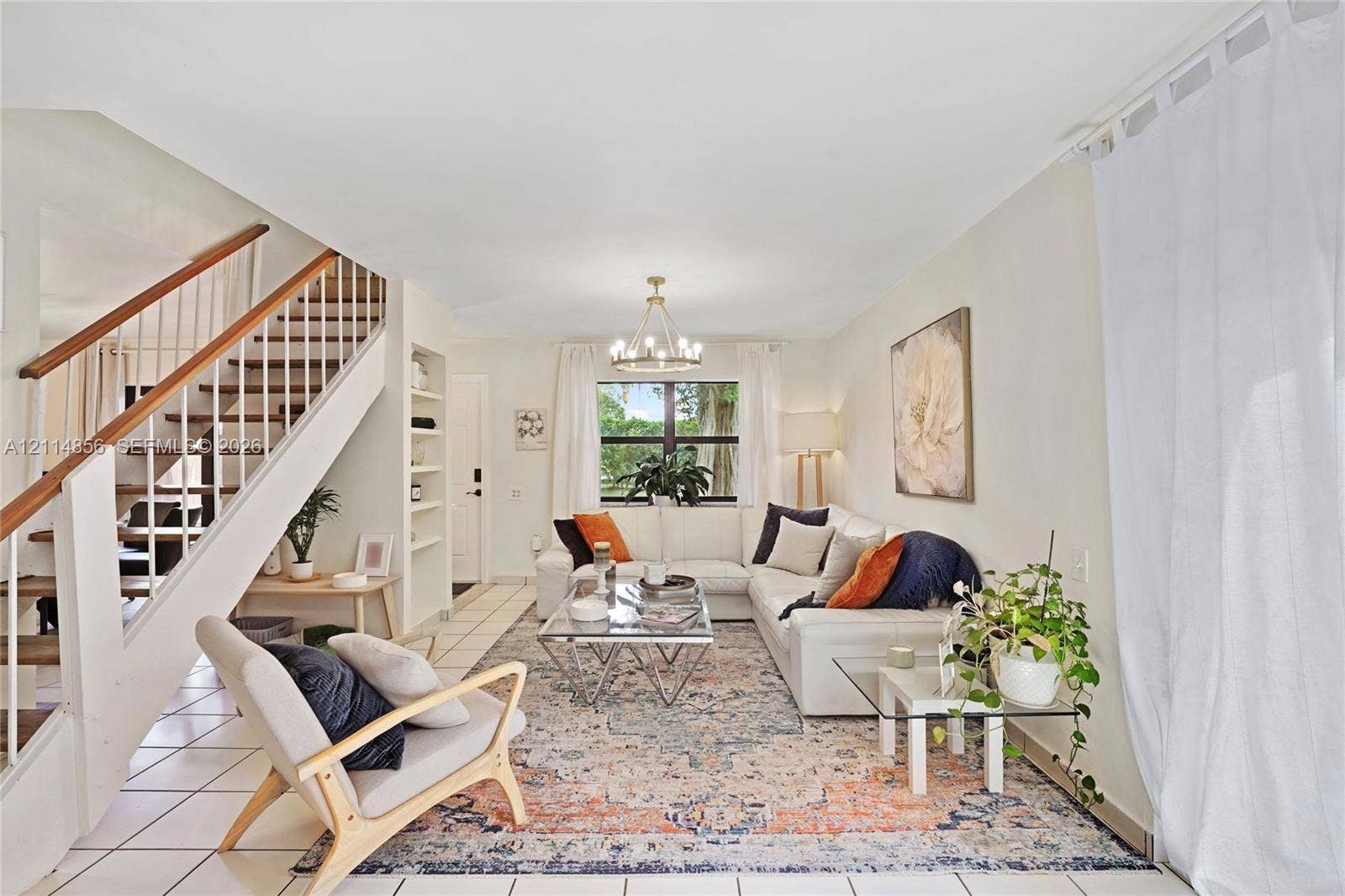 Miami, FL 33173 Open-concept living space featuring a central multi-arm chandelier, built-in recessed shelving, a staircase with wood-capped handrails, and tile flooring
