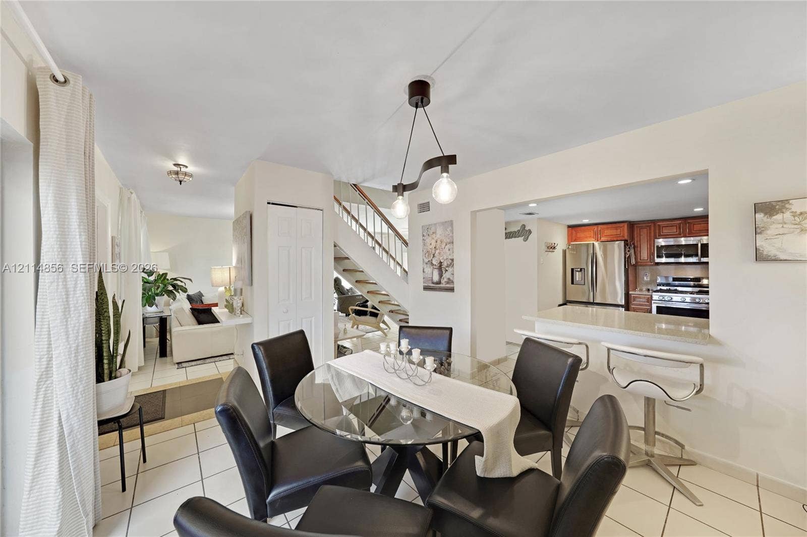 Miami, FL 33173 Open-concept interior featuring white ceramic tile flooring, a kitchen breakfast bar with light-toned solid surface countertops, wooden cabinetry, and a staircase with a wooden handrail